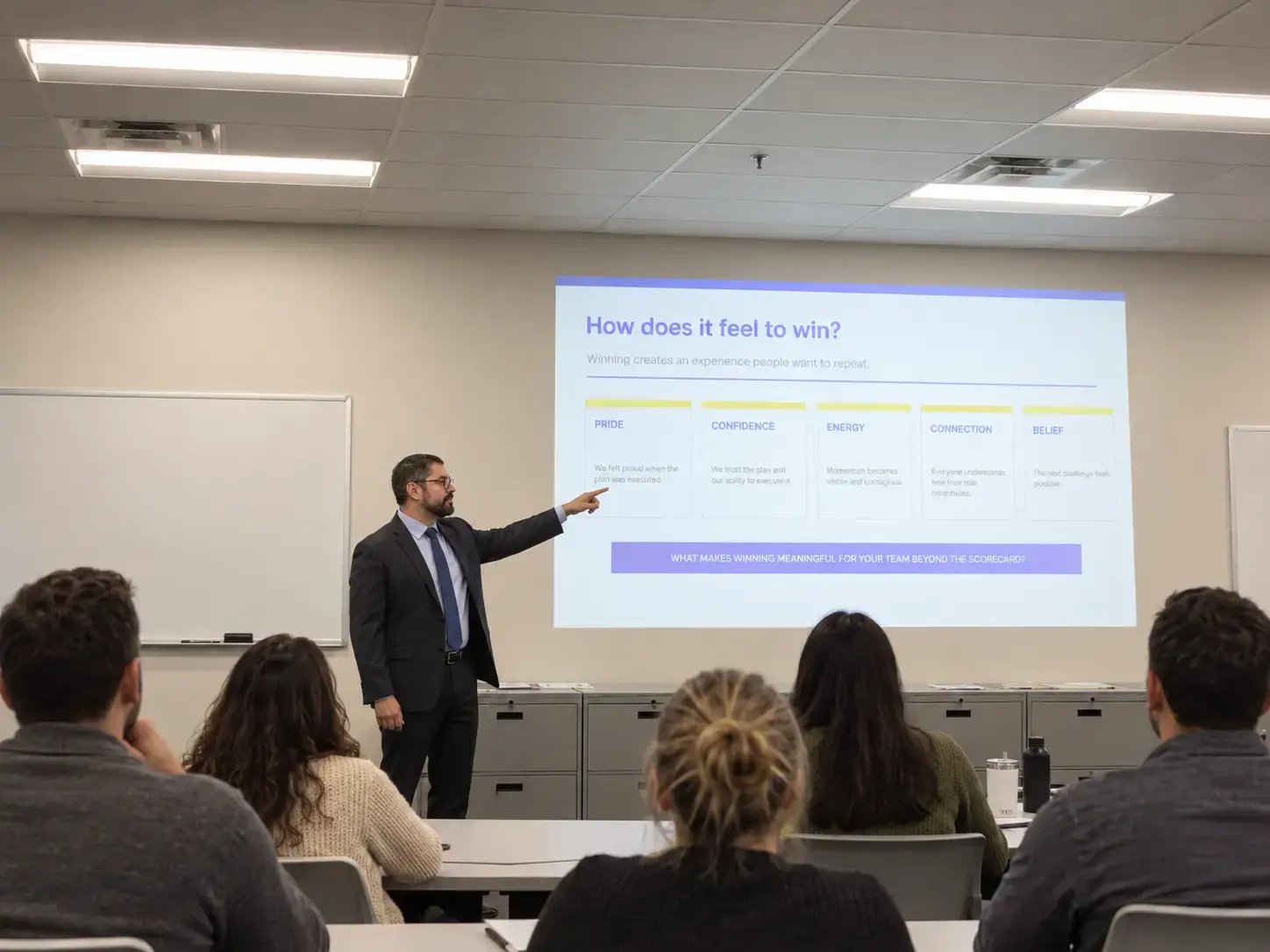 Presenter leading a corporate training session while pointing to a projected slide in front of seated attendees.